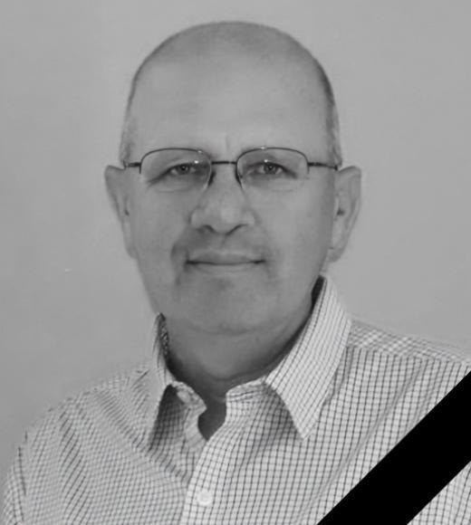 Black-and-white portrait of a Ken Tate smiling and wearing glasses and a checkered shirt, black memorial ribbon.